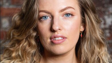 Portrait headshot of a woman with long wavy blonde hair and blue eyes, photographed against a textured brick wall. She is wearing a black top and small red earrings, with soft studio lighting and a natural makeup look. The image has a professional yet relaxed feel, with shallow depth of field keeping the focus on her face.