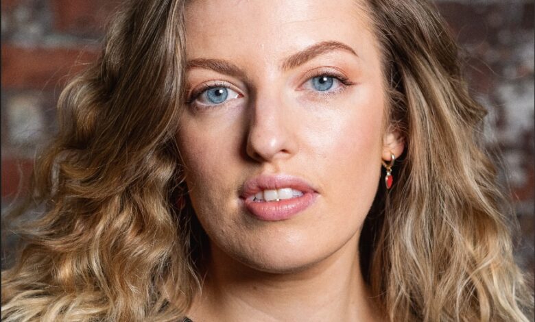 52 For 26 Poetry Project: Melissa Grindon 1 Portrait headshot of a woman with long wavy blonde hair and blue eyes, photographed against a textured brick wall. She is wearing a black top and small red earrings, with soft studio lighting and a natural makeup look. The image has a professional yet relaxed feel, with shallow depth of field keeping the focus on her face.