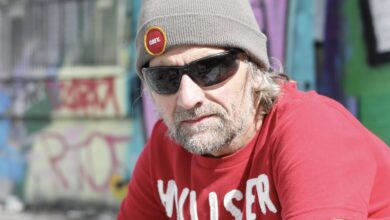 Portrait of a man wearing a grey beanie, black sunglasses and a red T-shirt, photographed outdoors in front of colourful graffiti-covered walls. He has facial hair and visible tattoos, with bright sunlight creating a candid urban street-style feel.
